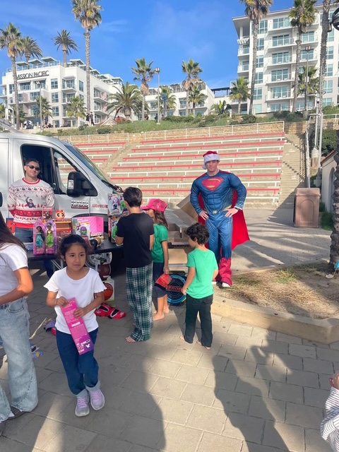 The Plumber Man in superhero suit giving gifts to kids at Oceanside Amphitheater